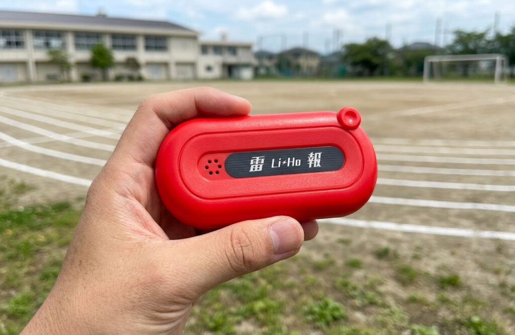 学校のグラウンドを背景に、男性の手が赤い長方形の雷検知器「雷 Li-Ho 報（らいほう）」を持っている。
