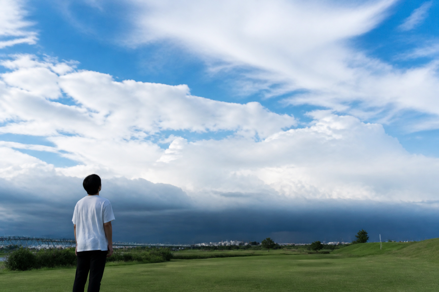 青空の向こうに黒い雲の帯が広がる河川敷で、空を見上げる人